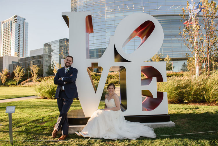 Bride and groom posing by a sculpture that spells "LOVE".