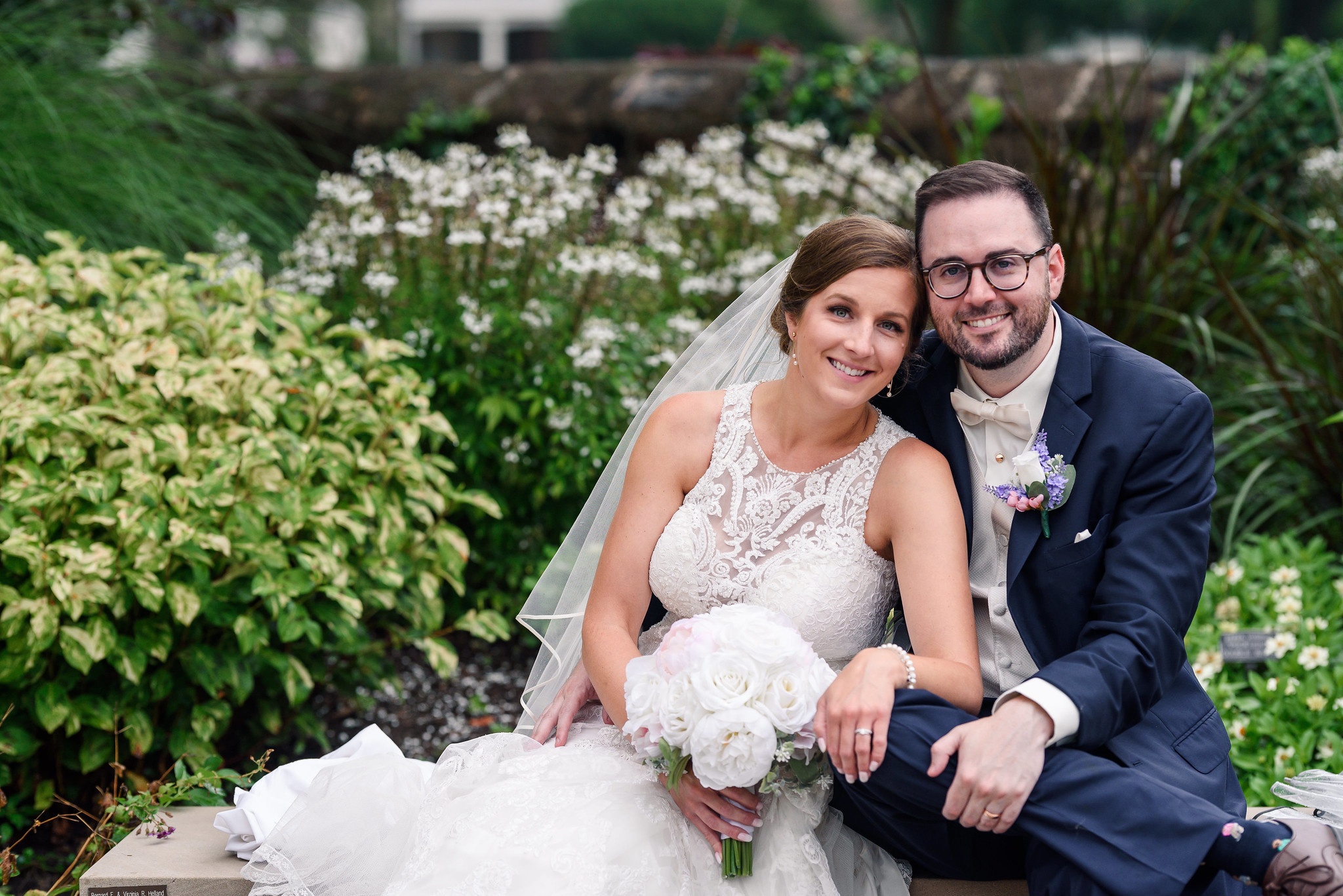 Bride and groom sitting together and smiling at the camera with pretty bushes in the background.
