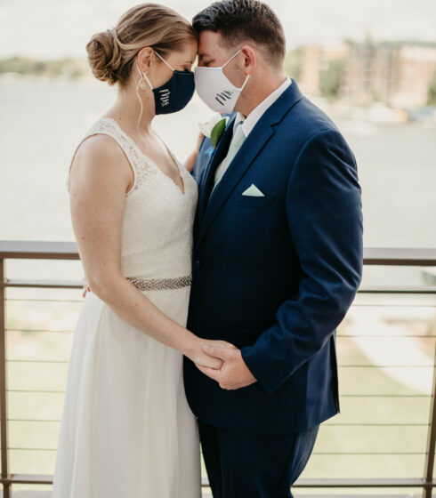 A bride and groom holding hands with their foreheads touching and their eyes closed while wearing masks labeled Mr. and Mrs.