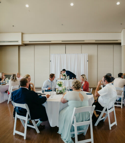 Wedding guest sitting at tables during reception with bride and groom kissing in the background.